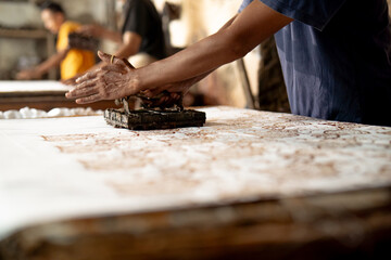 Close-up of Indonesian craftsman pressing copper stamp on fabric to create batik pattern in traditional workshop, handmade textile art from Java.