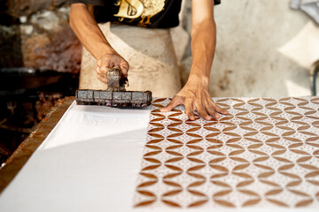BATIK CAP - Traditional craftsman printing batik pattern using copper stamp on white cloth at local workshop in Pekalongan, Indonesia, representing cultural heritage and handmade textile art.