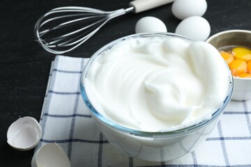 Whipped egg whites in bowl, whisk and ingredients on black table, closeup