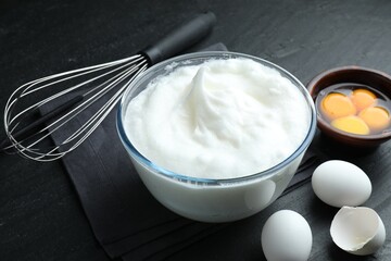 Whipped egg whites in bowl, whisk and ingredients on black table, closeup