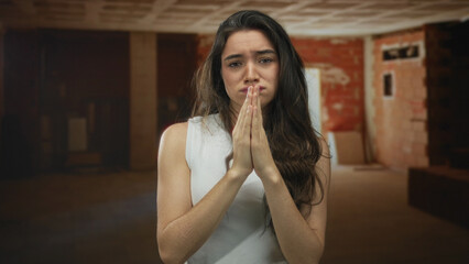 Young hispanic woman pressing hands in prayer at construction site interior; desperate plea for relief.