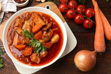 Delicious cooked stew in bowl and fresh ingredients on wooden table, flat lay