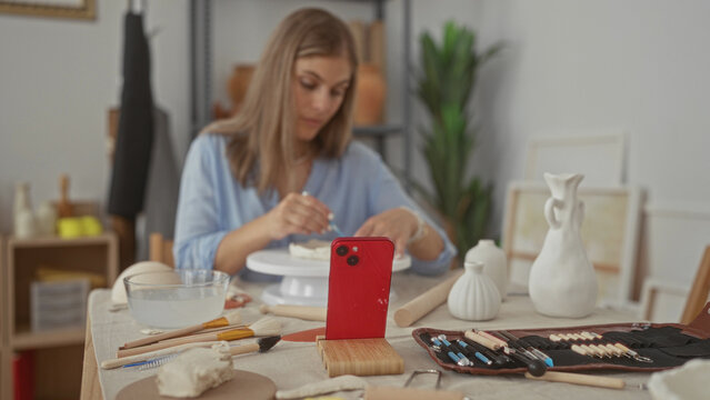 Woman sculpts clay with tool in artisan studio surrounded by pottery brushes and wheel on table; concentration.