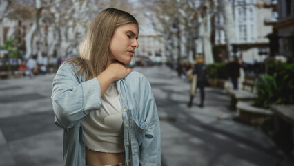Fototapeta premium Woman holding neck in pain on busy city street with blurred pedestrians, benches, and bare trees; discomfort.