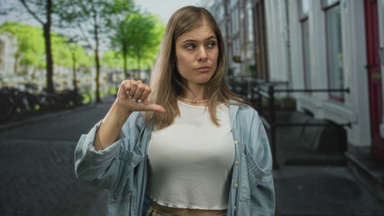 Fototapeta premium Blonde woman in cropped white top shows thumbs down gesture on city street lined with bicycles and brick buildings; rejection.