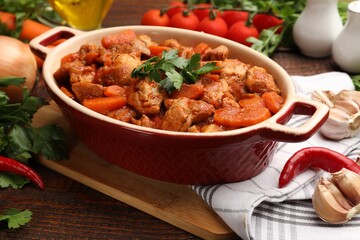 Delicious cooked stew in baking dish and fresh ingredients on wooden table, closeup