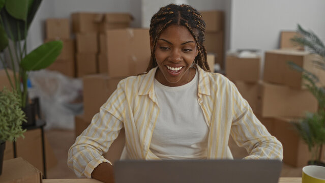 Woman at laptop wearing striped shirt with hand to chest, surrounded by moving boxes in building; new home excitement.