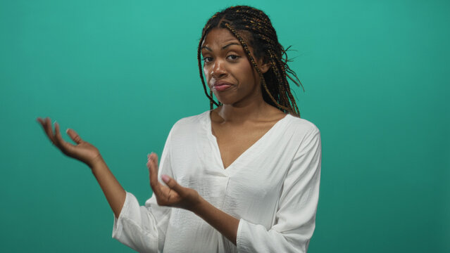 African american woman in white blouse presenting open palms toward camera in teal studio with braided hair; skeptical curiosity.