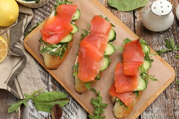 Tasty bruschettas with salmon, cucumber and arugula served on wooden table, flat lay