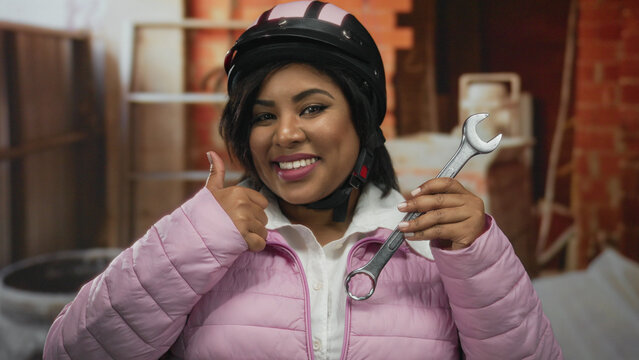 Young latin woman wearing helmet holding wrench indoors at construction site showing positive attitude with smile and thumbs up in pink jacket.