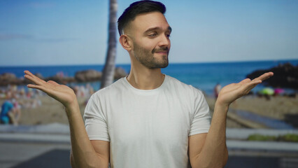 Young hispanic man in white shirt making a shrugging gesture on a sunny beach with blue sky and ocean in the background showing a casual and relaxed vibe outdoors.