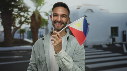 Young man smiling while holding a philippine flag on an urban city street, showcasing cultural pride in a vibrant cityscape background outdoors.