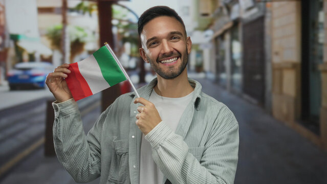 Fototapeta Young man smiles holding an italian flag on a city street, capturing a vibrant urban scene outdoors with cultural pride.