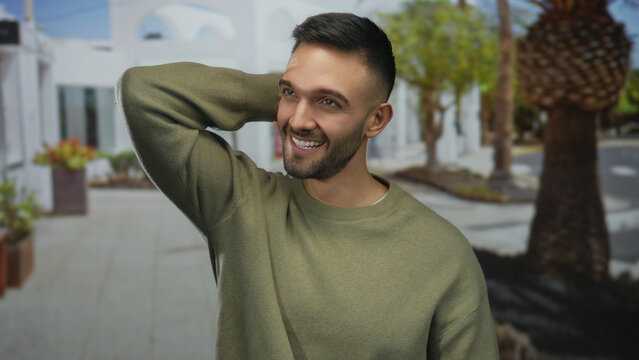 Young man smiling outdoors on a sunny day in an old urban town street, embracing the vibrant atmosphere with a joyful expression against a backdrop of rustic architecture and greenery.