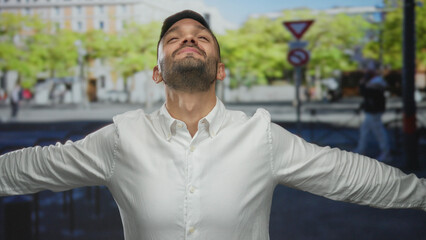 Young man in white shirt stands joyfully in an urban street setting with arms wide open, expressing...