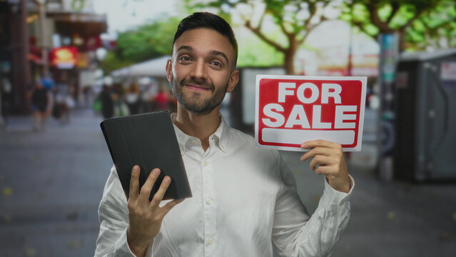 Hispanic man smiling with tablet and for sale sign standing outdoors in bustling urban street