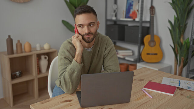 Young man talking on phone in modern living room, sitting at desk with laptop, chart, and notebook, conveying a relaxed indoor work environment.