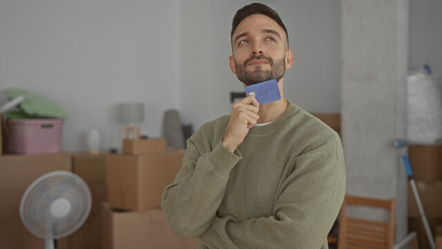 Young man indoors in new home holding a blue credit card surrounded by moving boxes looking thoughtful in a casual environment with cardboard and minimal furniture around him