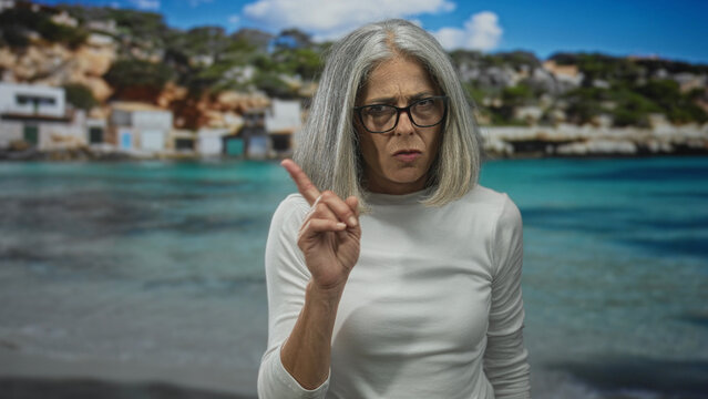 Woman finger to lips for silence on beach with grey hair and eyeglasses near seaside houses, white shirt visible; serious warning. - Powered by Adobe