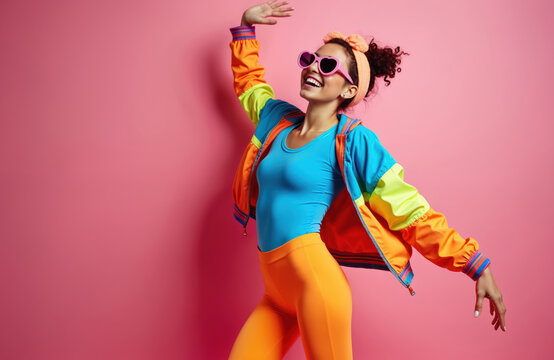 Young woman in vibrant 80s fashion outfit dances with joy. Wears colorful jacket, orange leggings, pink heart sunglasses. Happy female model poses on pink studio background, showing retro style. Girl