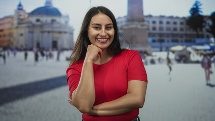 Woman with hand on chin and crossed arms in front of a historic building plaza wearing a red tshirt, smiling and posing naturally; confidence.