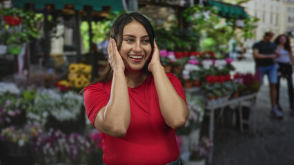 Woman covering mouth with hands at flower stall on street market, hands to face gesture and smiling; surprise candid moment.