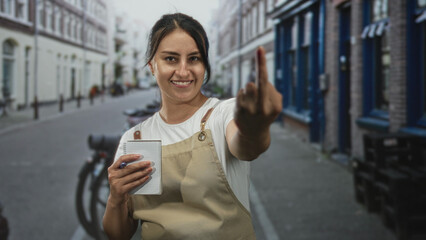 Woman waitress in beige apron holds notepad and pen and shows middle finger while smiling on city street; defiance.