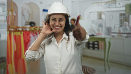 Young woman architect wearing white hardhat cups hand to ear while leaning toward colorful clothing racks in a retail building, listening for distant noise; curiosity.
