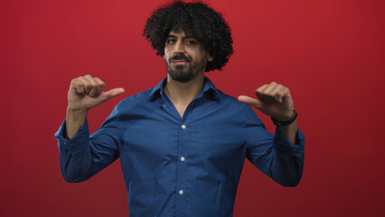 Man with curly hair and beard, thumbs pointing to chest, wearing blue shirt in red studio; confidence.