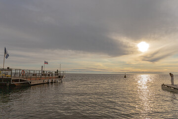 Quiet harbor as a wooden pier extends into the sea. Flags flutter, a lone figure stands on the dock, and the water mirrors a tranquil sky.