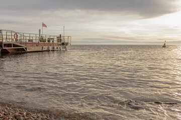 A quiet seascape with a wooden pier extending into calm water at sunset. Pebble shoreline, distant buoy, and a waving Russian flag.