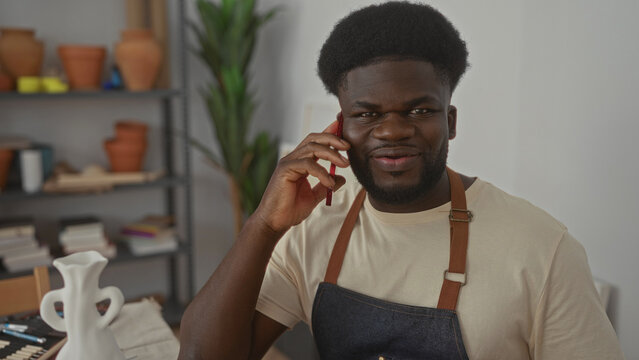 Man holds smartphone to ear and smiles while wearing apron in studio surrounded by pottery and tools; craftsmanship contentment. - Powered by Adobe