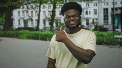 Fototapeta premium Man showing thumbs up on street in front of white apartment building and green park hedges, smiling with bare hand and thumb visible; confidence positivity.