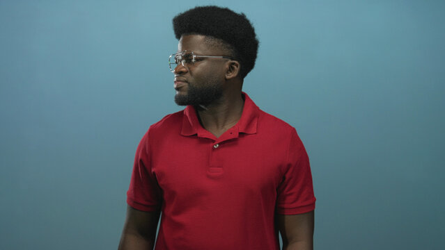 Young african american man in red polo with glasses turns head left against blue studio wall; contemplation poise. - Powered by Adobe