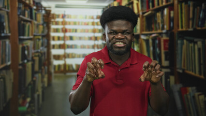 Man snarling and clawing with hands in a library building, wearing red polo shirt and showing teeth while posing between bookshelves; playful mischief.