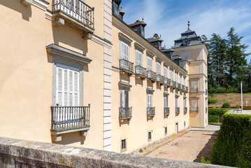 The classic facade of the Royal Palace of El Pardo, featuring its ochre walls, steep slate roofs, corner tower, and the adjacent chapel structure under a bright blue sky © alanfalcony