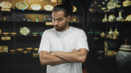 Man with arms crossed showing forearms in museum gallery wearing white tshirt and beard; skepticism curiosity doubt.