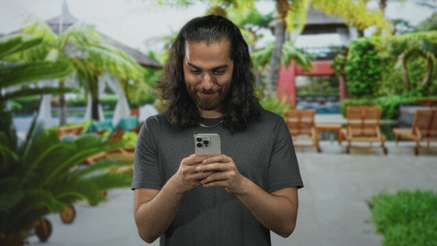 Man smiling and holding smartphone with visible hands and braces while looking down at screen at resort building poolside; relaxed vacation happiness.
