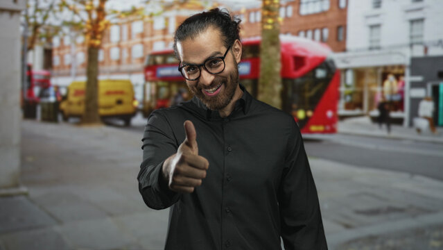Man giving a thumbs up gesture on a city street with red double decker bus and delivery van nearby, smiling and wearing glasses; approval confidence friendliness.