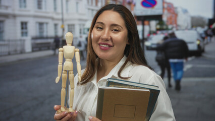 Woman holds wooden mannequin and notebooks on a city street, smiling at camera and displaying model figure in hand; creativity.
