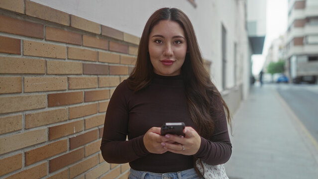 Hispanic woman scrolls smartphone on city street by brick wall while reading messages; calm contentment.