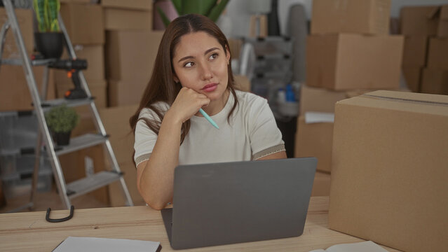 Hispanic woman with laptop and pen rests chin on hand among stacked moving boxes in building; contemplation.