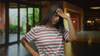 Young hispanic woman covering her face with hand and glasses while standing in a hotel building entrance hall; embarrassment.