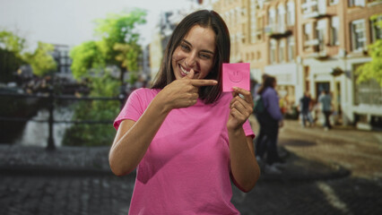 Fototapeta premium Woman points finger to a pink sticky note with a drawn smile, wearing a pink tshirt and holding it near her face on a street in amsterdam; happiness.