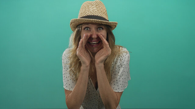 Woman cupping hands around mouth and calling out in studio with teal backdrop, wearing straw hat and polka dot dress; playful secret.