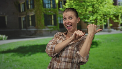 Fototapeta premium Woman smiling and points thumb to building entrance on a green lawn wearing a plaid shirt and rolled sleeves while looking aside; joy welcome invitation.