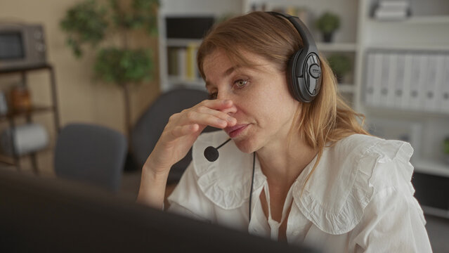 Woman with headset microphone holding forehead and hand to temple at office desk, headset mic visible and gesture suggests headache; work stress.