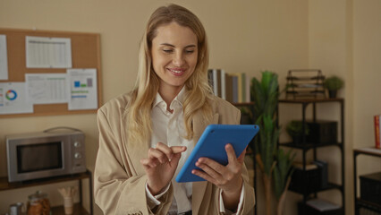 Woman using tablet in modern office setting with a smile, surrounded by business charts on bulletin board and office supplies, conveying professional and productive atmosphere.