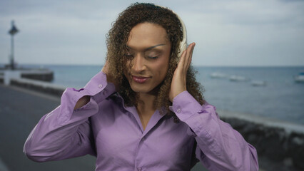 Transgender woman holding headphones with eyes closed listening to music on street; serenity and empowerment.