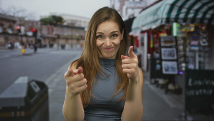 Young caucasian woman pointing both fingers directly at camera on lively city street; playful confidence.